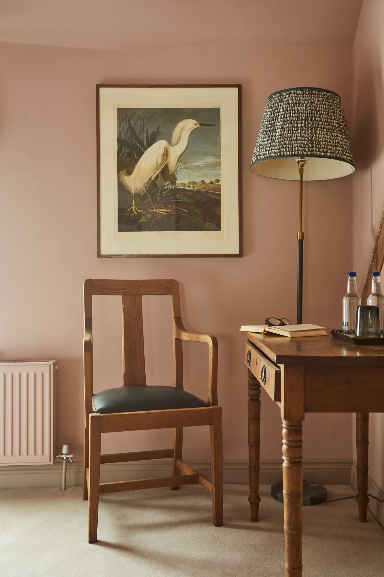 A corner of a pink bedroom with an egret print, an antique writing desk and chair, and a checked lampshade