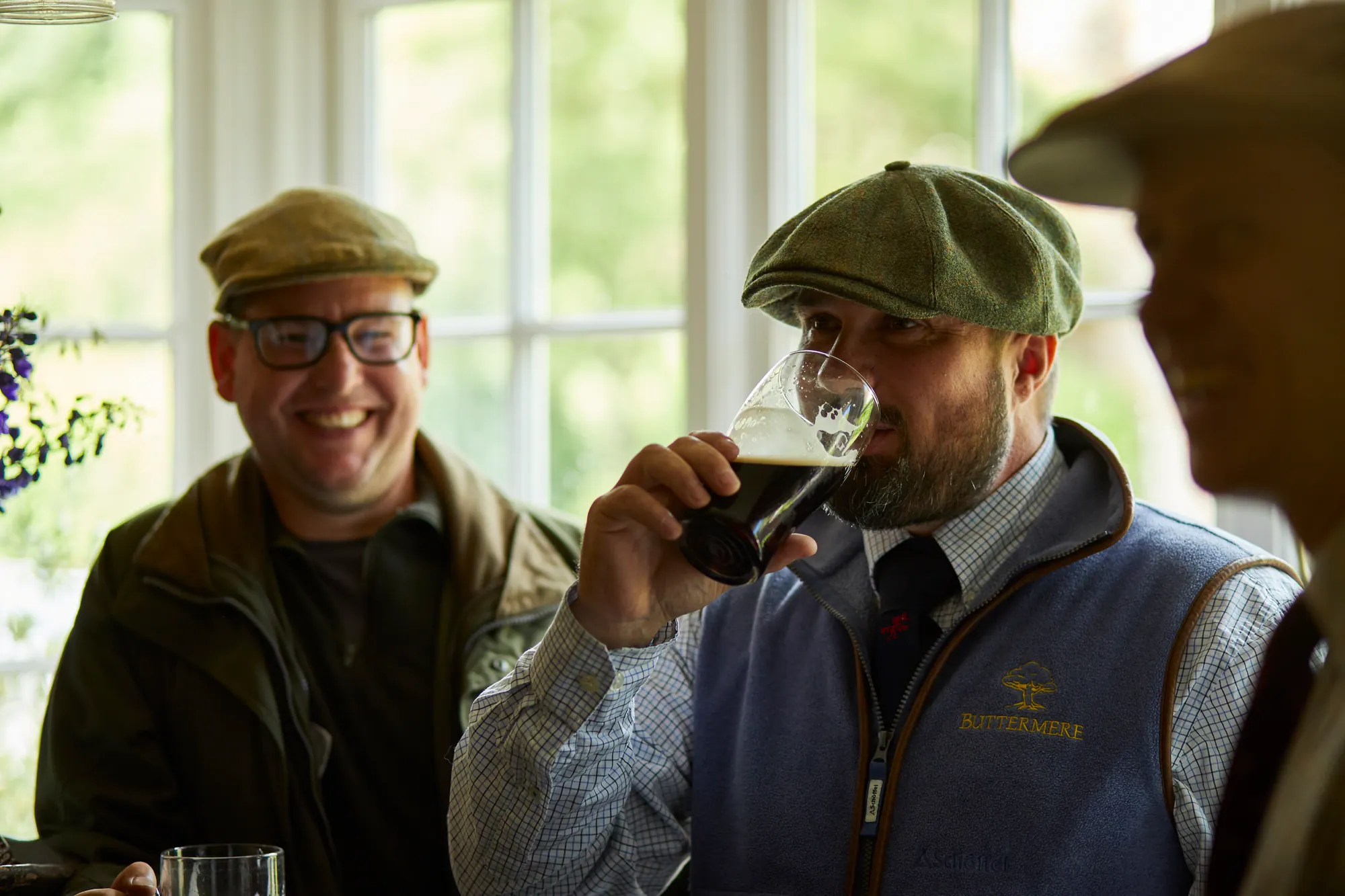 Two friends in flat caps by the window, one laughing, the other taking a sip of stout