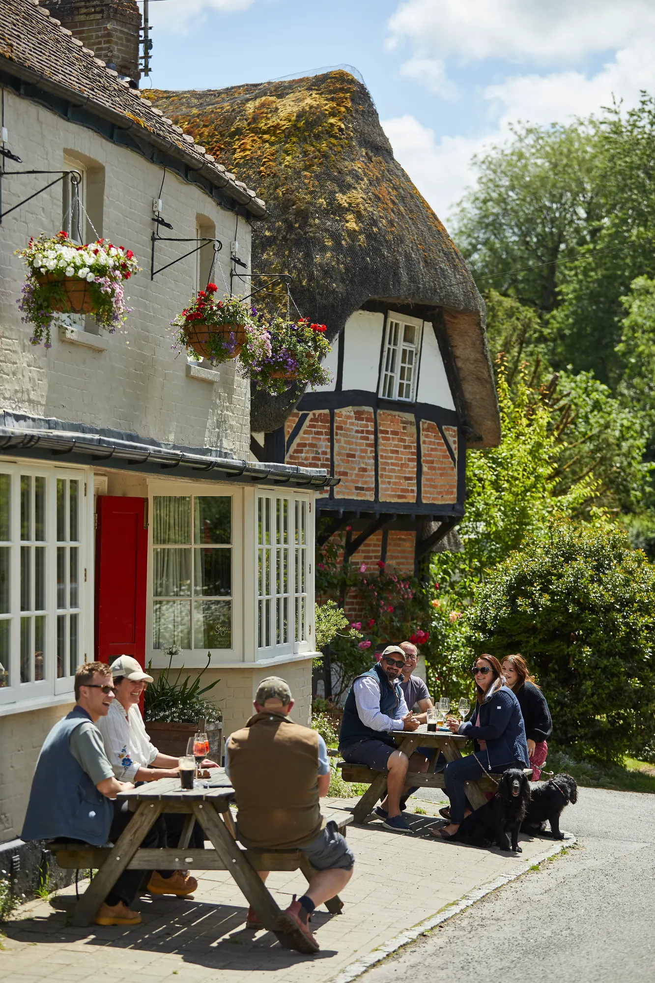Guests and their dogs at picnic tables outside the pub on a sunny afternoon, the thatched cottage next door behind
