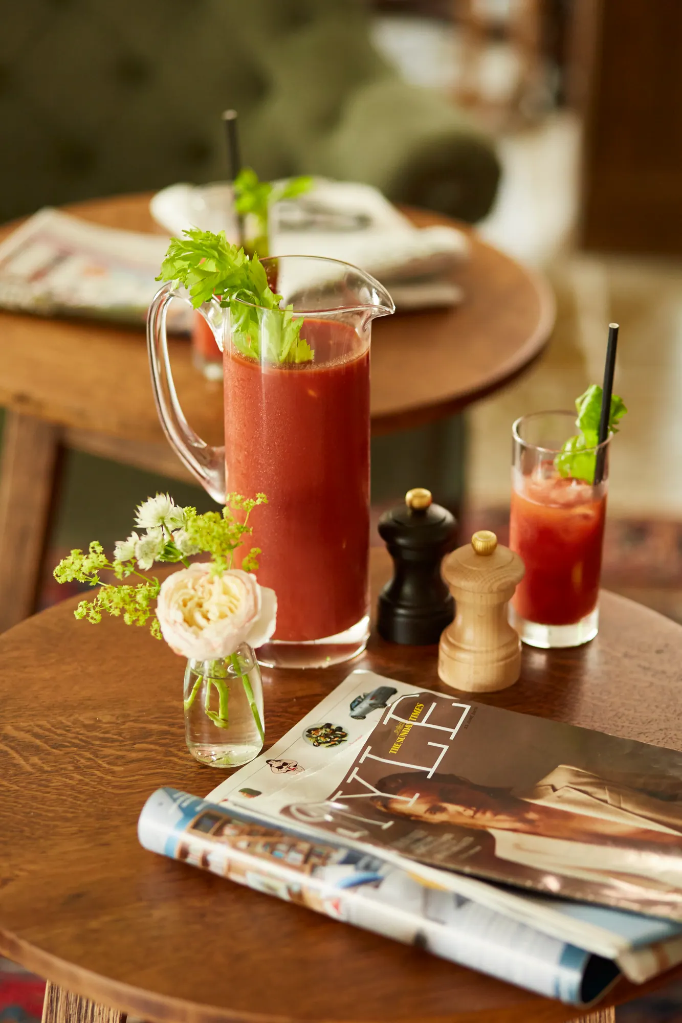 A jug and glass of Bloody Mary with celery sticks, beside fresh flowers and the Sunday papers