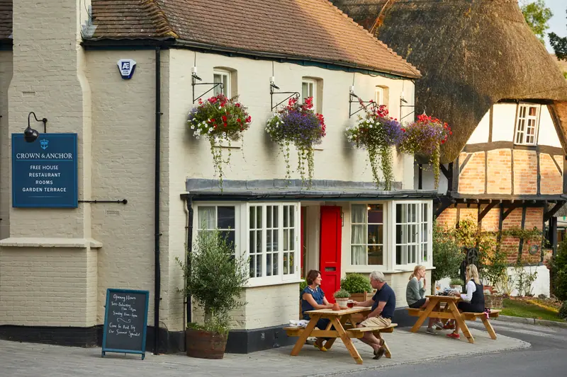 The cream-painted front of the Crown & Anchor in Ham, hanging baskets in bloom and guests at picnic benches by the red door