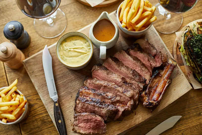 A sliced steak on a carving board with chips, béarnaise, a jug of gravy and glasses of red wine