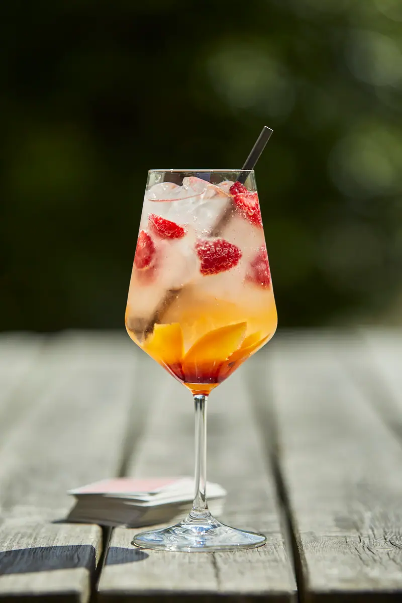 A spritz with raspberries and a slice of orange in a stemmed glass, on a weathered table outside