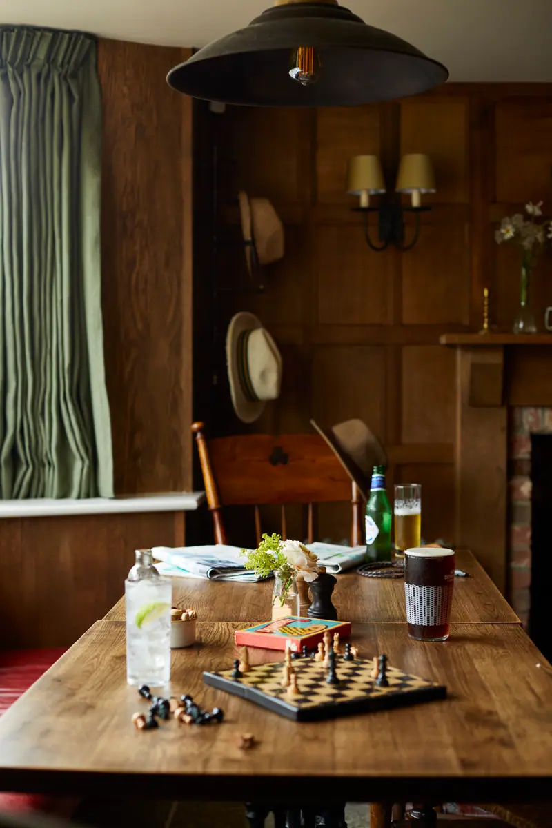 A corner table set with a chess game and a drink, felt hats hanging on the panelled wall behind