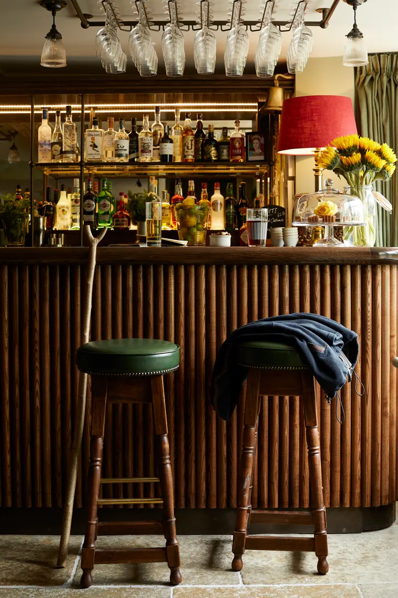 The bar, with a reeded wooden front, green leather stools, a red lampshade and sunflowers among the bottles