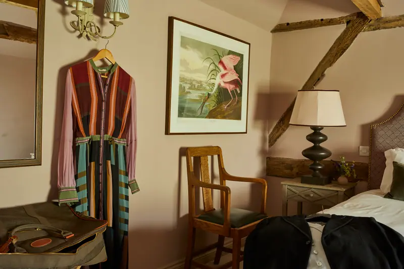 A pink bedroom under old beams, with an Audubon flamingo print, a striped dressing gown on the wall and an antique chair