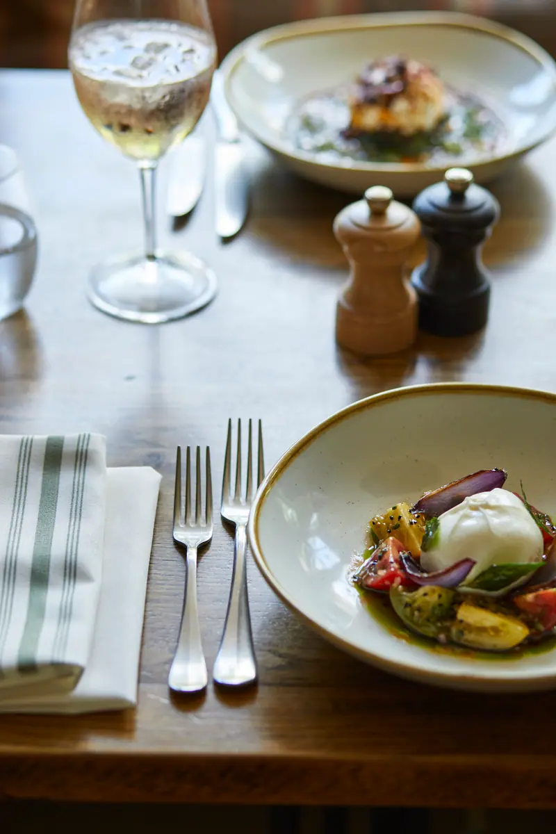 Burrata with heritage tomatoes and red onion, a glass of white wine and a second plate across the table