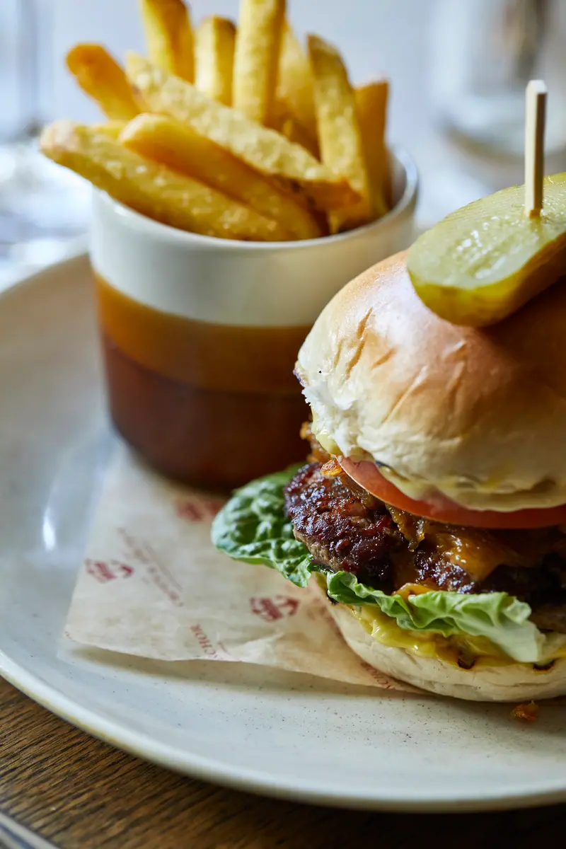 A cheeseburger topped with a gherkin, with a pot of chips