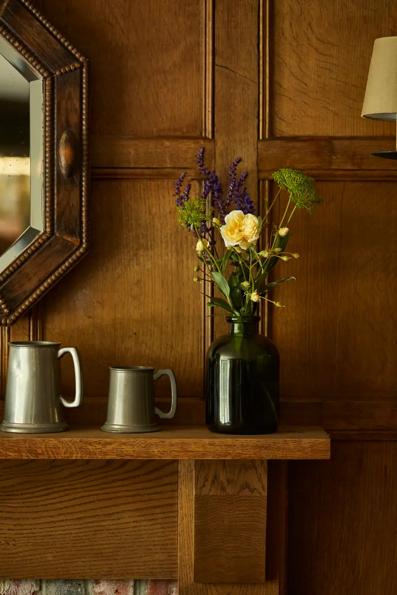 Garden flowers in a dark glass vase beside pewter tankards on a panelled shelf