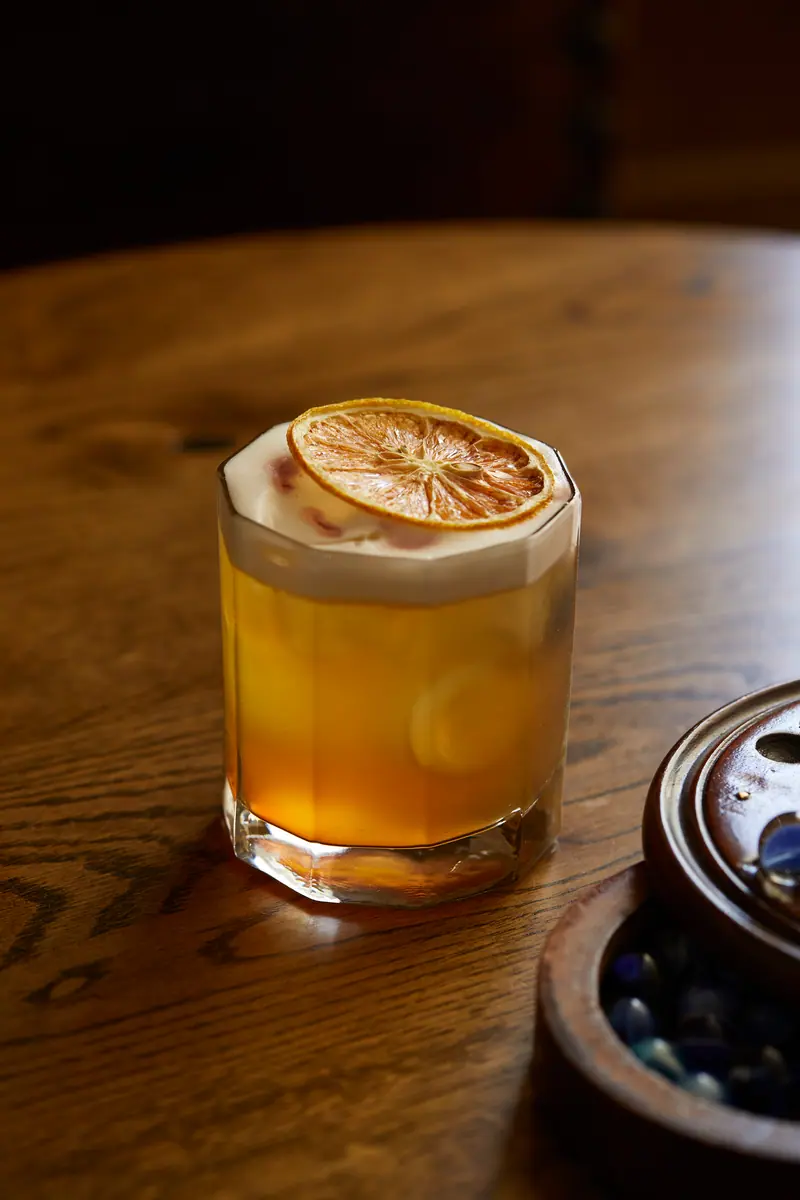 A whisky sour with a frothy top and a dried orange wheel, on a round oak table