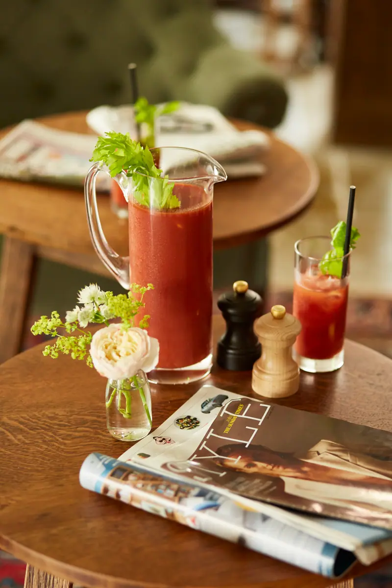 A jug and glass of Bloody Mary with celery sticks, beside fresh flowers and the Sunday papers